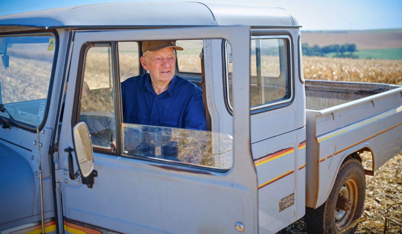 agricultor idoso Adolfo Midding, de boné e camisa azul, olhando para fora pela janela do motorista de sua caminhonete Toyota Bandeirante antiga, estacionada ao lado de uma lavoura de milho safrinha.